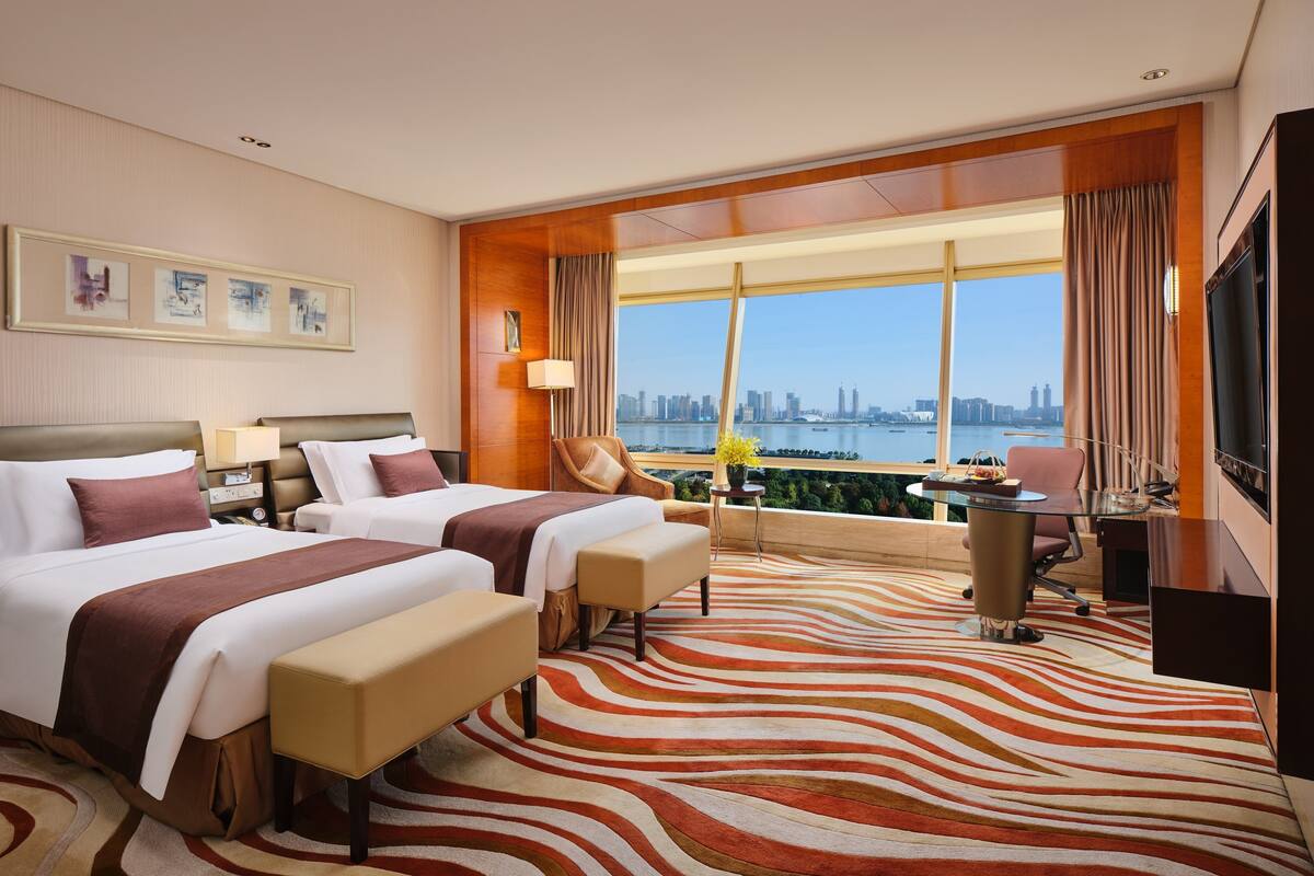 A hotel room with two beds, a desk, a chair, a TV, and a large window with a city view.