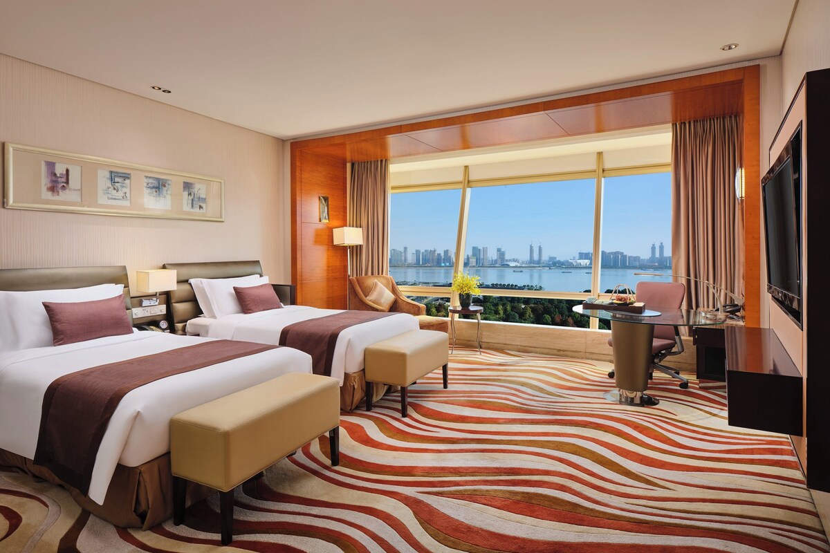 A hotel room with two beds, a desk, a chair, a TV, and a large window with a city view.