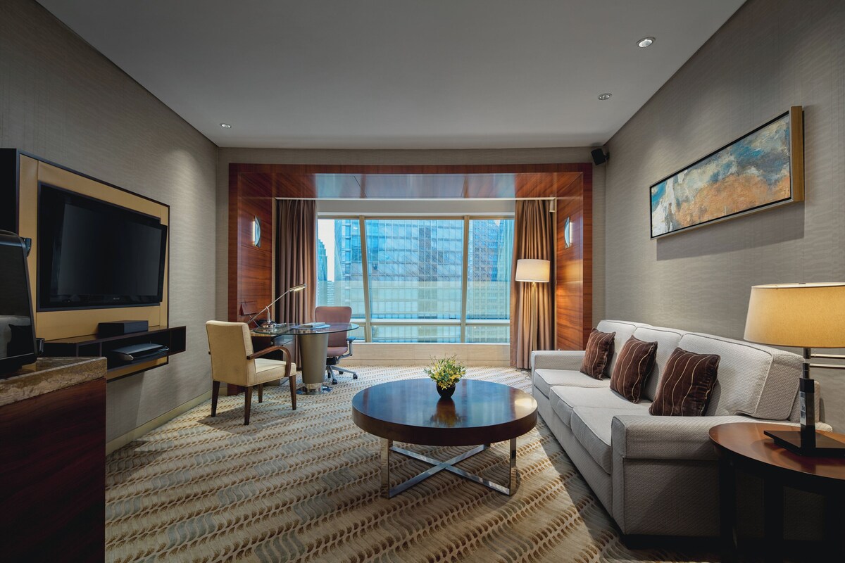 A modern hotel room with a large flat-screen TV, a round coffee table with a plant, a sofa with cushions, and a large window offering a city view.