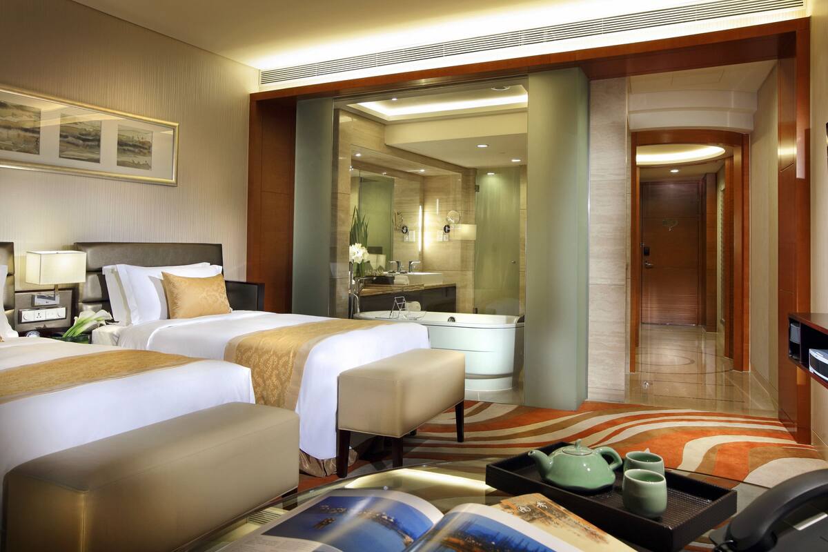 A hotel room with two beds, a sitting area, a bathroom with a mirror, and a TV.