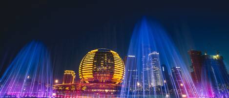 A nighttime cityscape with illuminated buildings and a water fountain display.