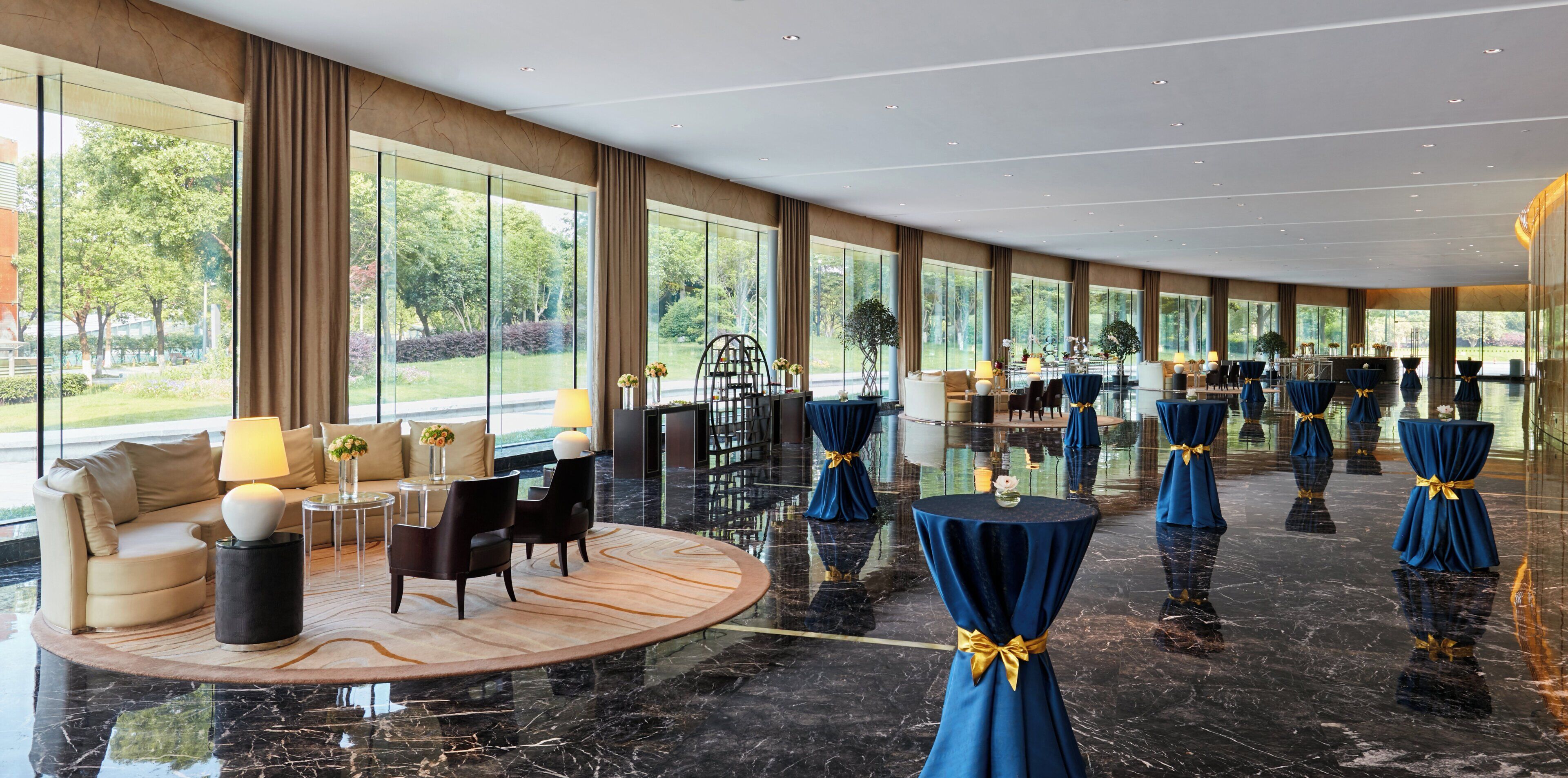 Un amplio vestíbulo de hotel con piso de mármol, ventanas grandes que ofrecen vistas al exterior y un área de descanso con sofá y sillones.