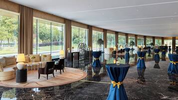 Un amplio vestíbulo de hotel con piso de mármol, ventanas grandes que ofrecen vistas al exterior y un área de descanso con sofá y sillones.