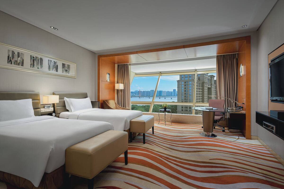 A hotel room with two beds, a desk, a chair, a TV, and a large window with a city view.