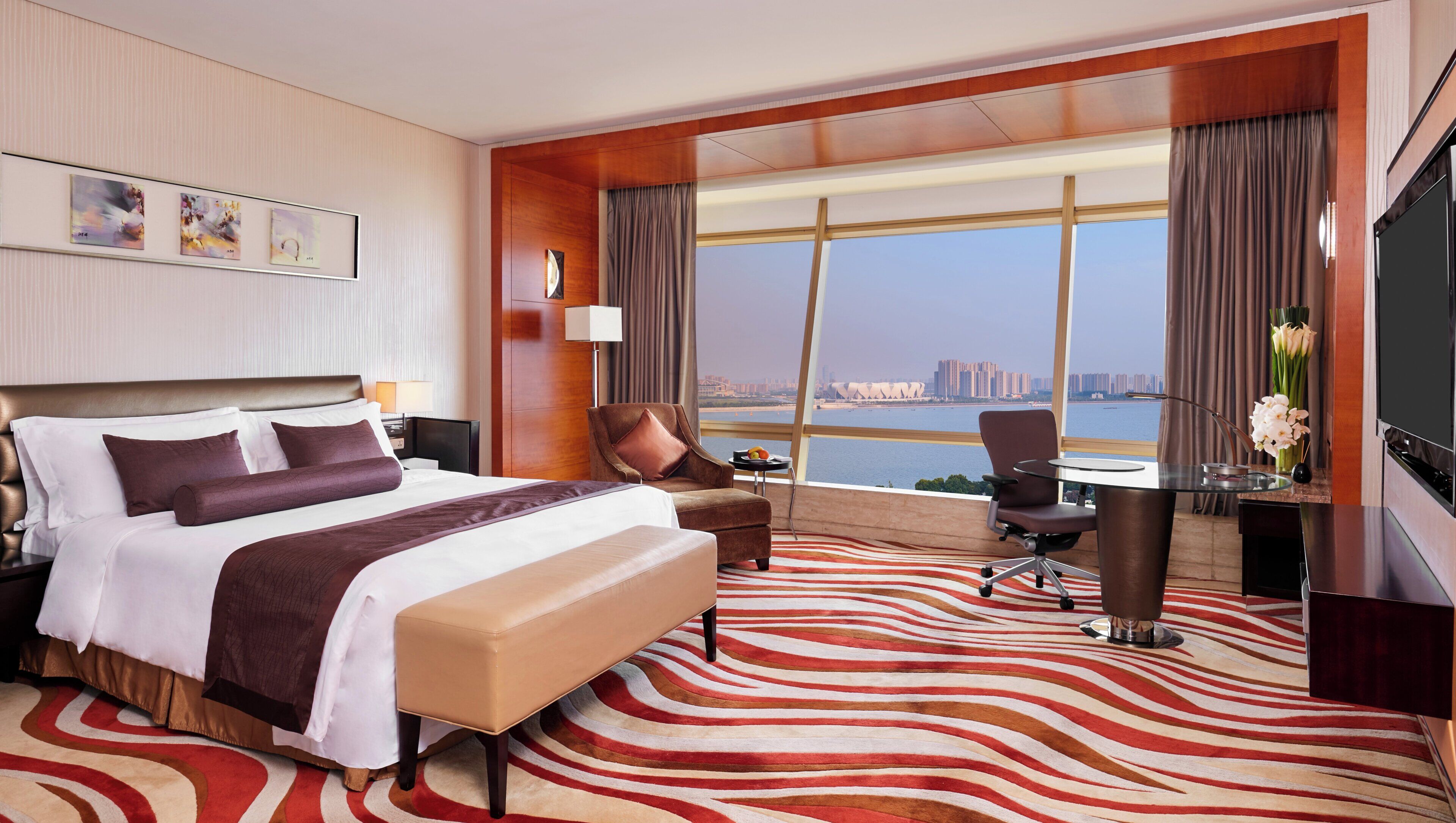 A hotel room with a large bed, a desk, a chair, a TV, and a view of a cityscape.