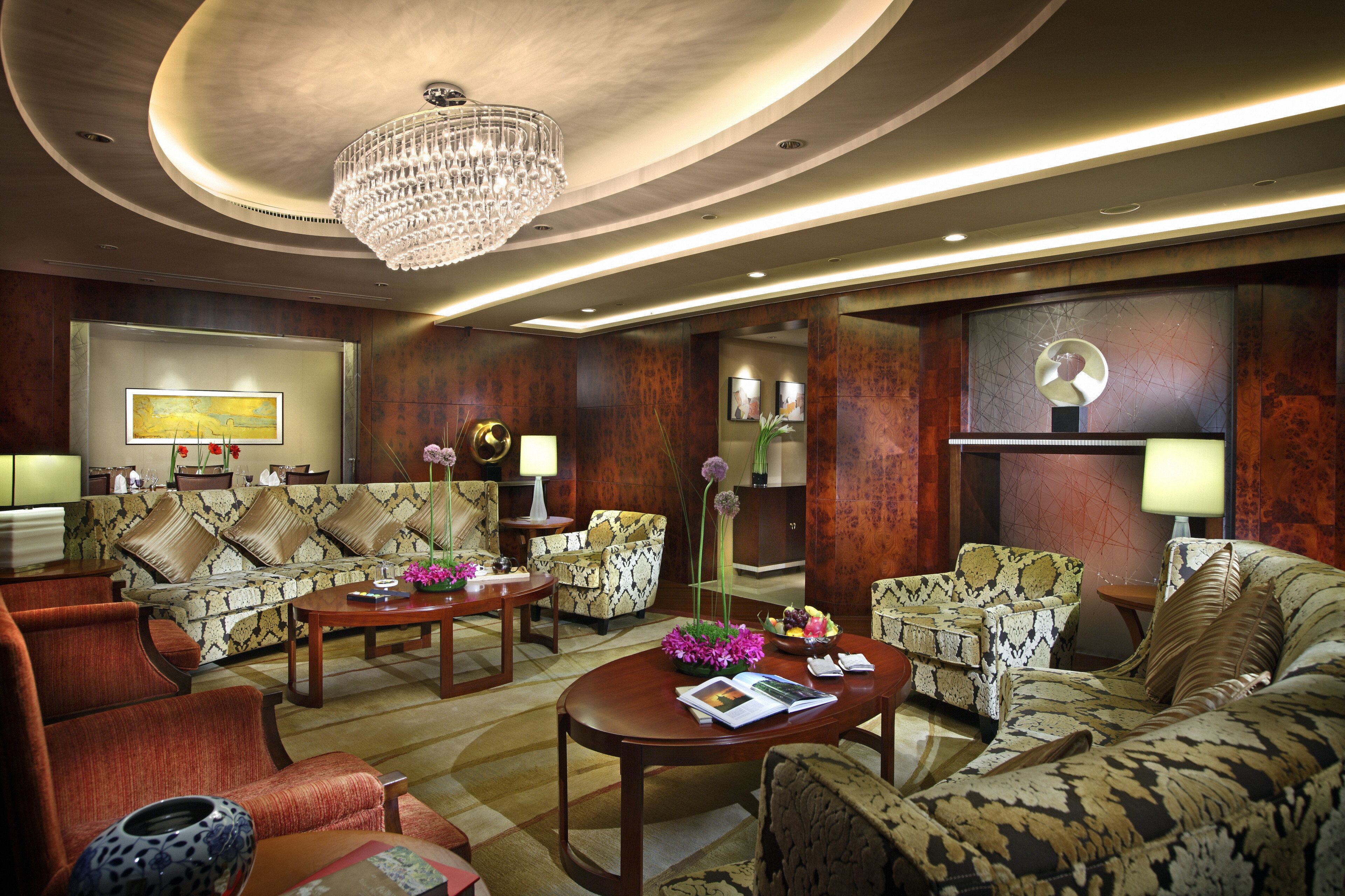 A hotel lobby with a chandelier, patterned sofas, a coffee table, and a dining area.