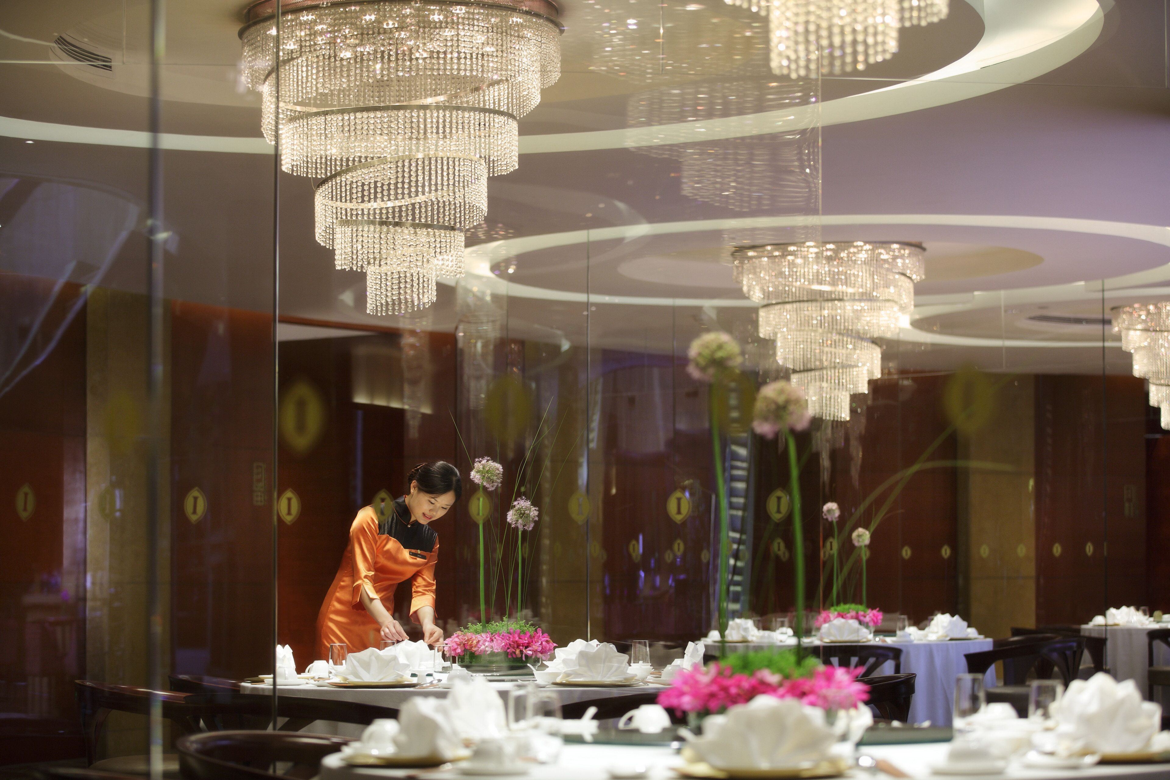 L’intérieur d’un restaurant, avec plusieurs tables dressées pour le service, un membre du personnel s’occupant d’une table et un grand lustre.