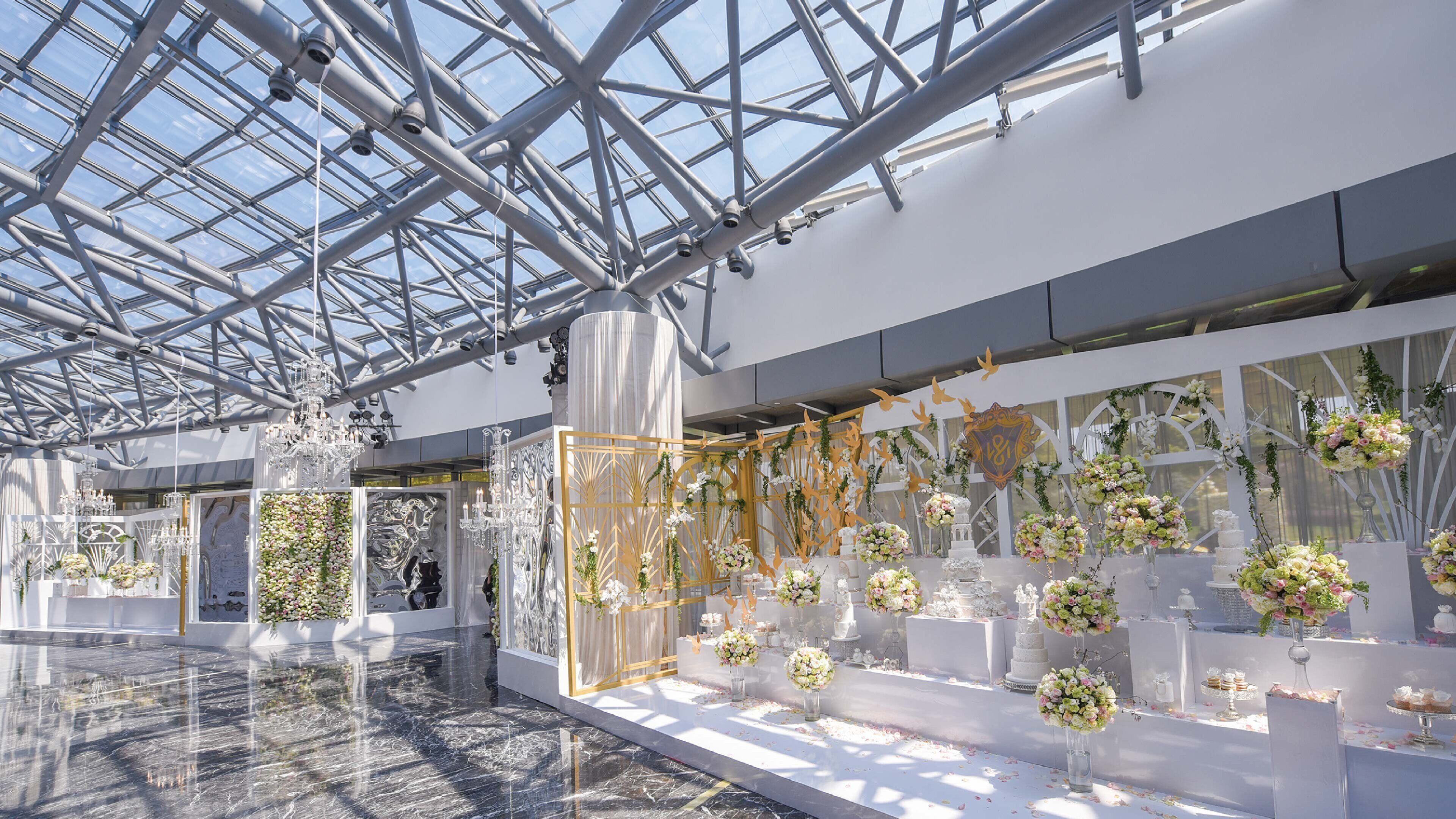 A spacious indoor area with a high ceiling and a glass roof, featuring a floral arrangement and a white reception desk.