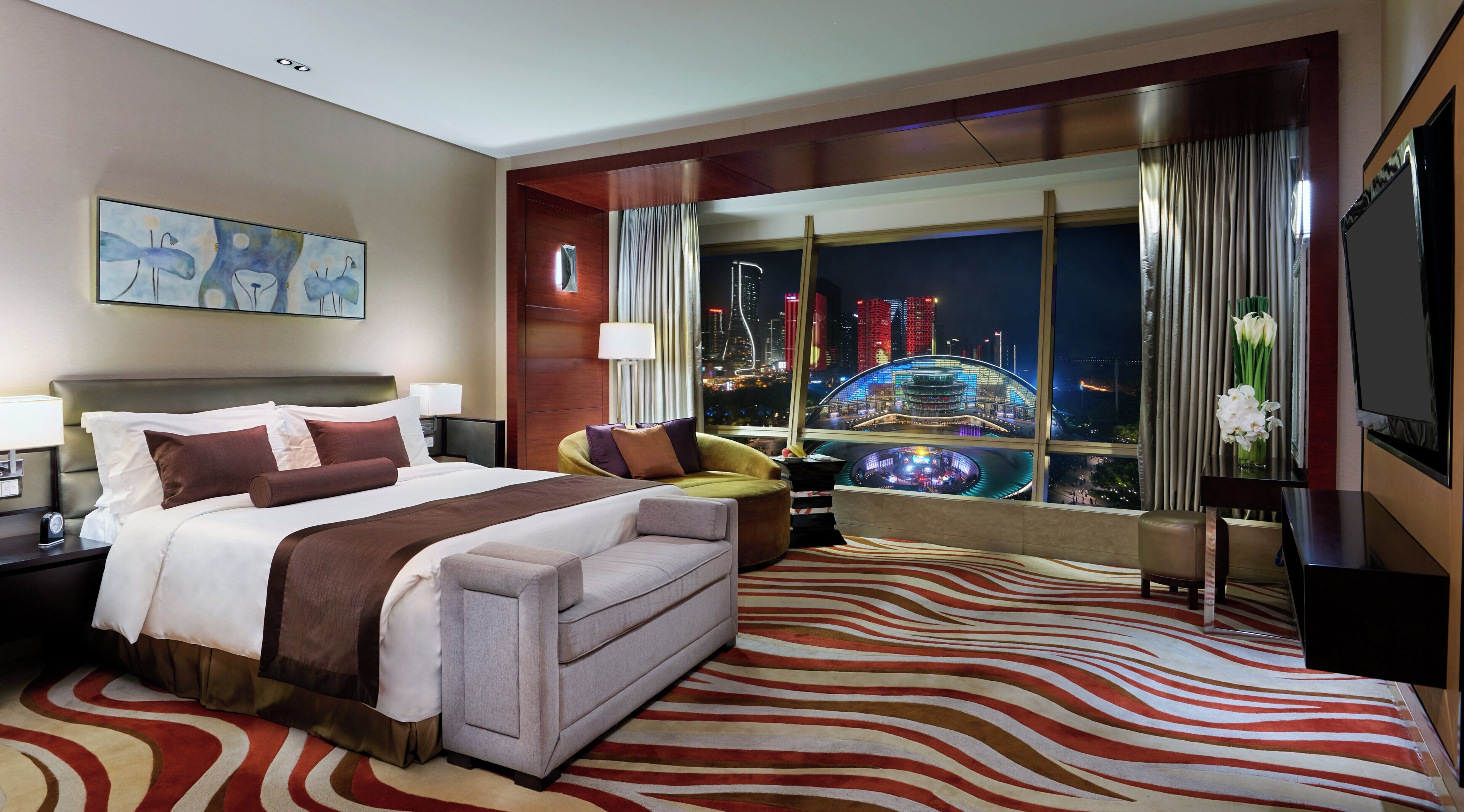 A hotel room with a bed, a sofa, a view of a cityscape, and a large window with curtains.