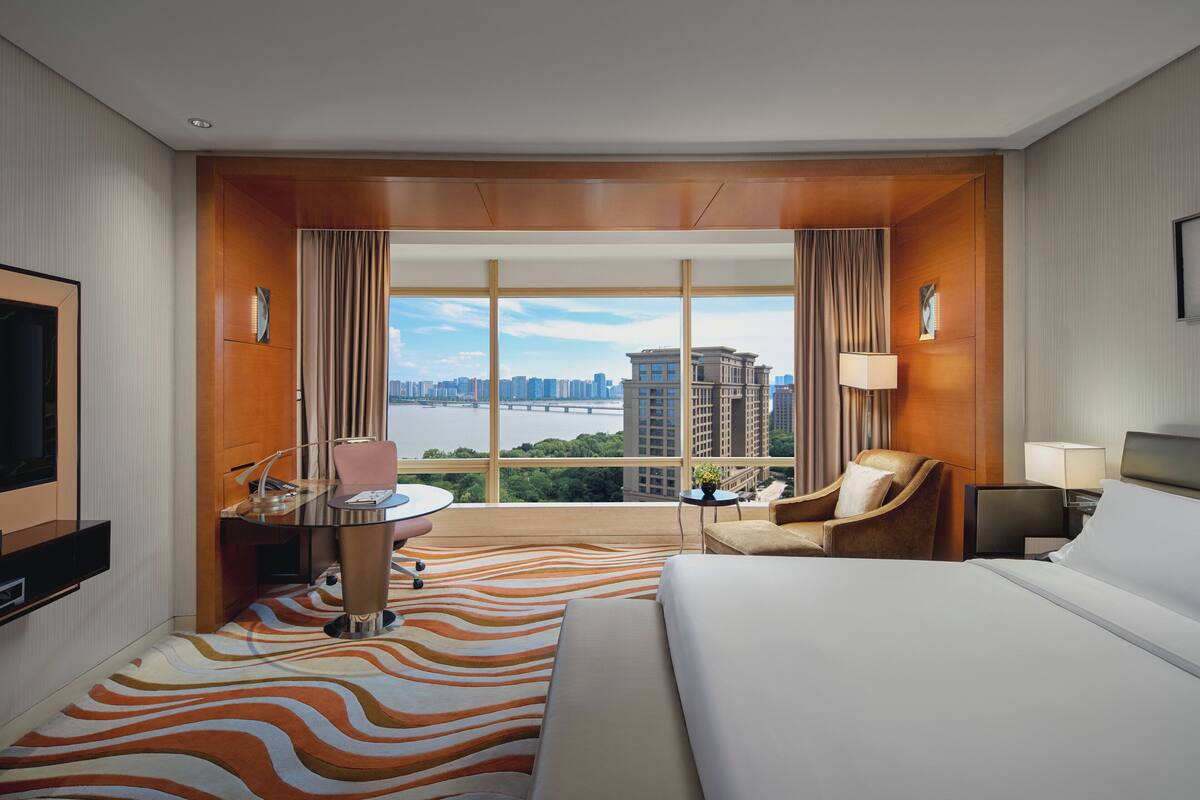 A hotel room with a large bed, a desk with a chair, a TV, and a view of a cityscape and river.