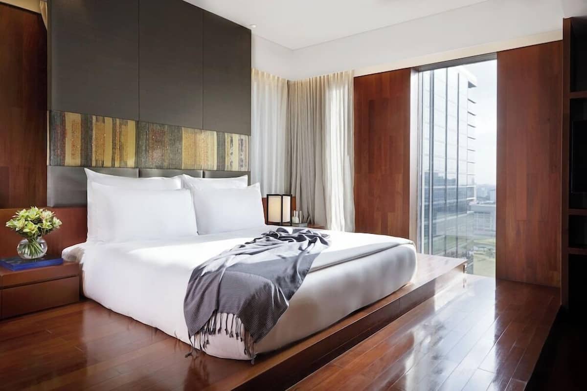 A modern hotel room with a large bed, a nightstand with a vase of flowers, and a view of a cityscape through large windows.