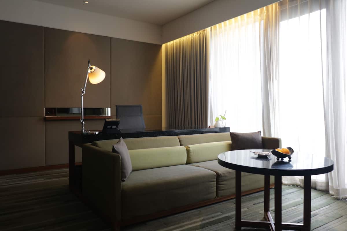 A modern hotel room with a sofa, a desk with a laptop, a round table with a bowl, and a lamp.