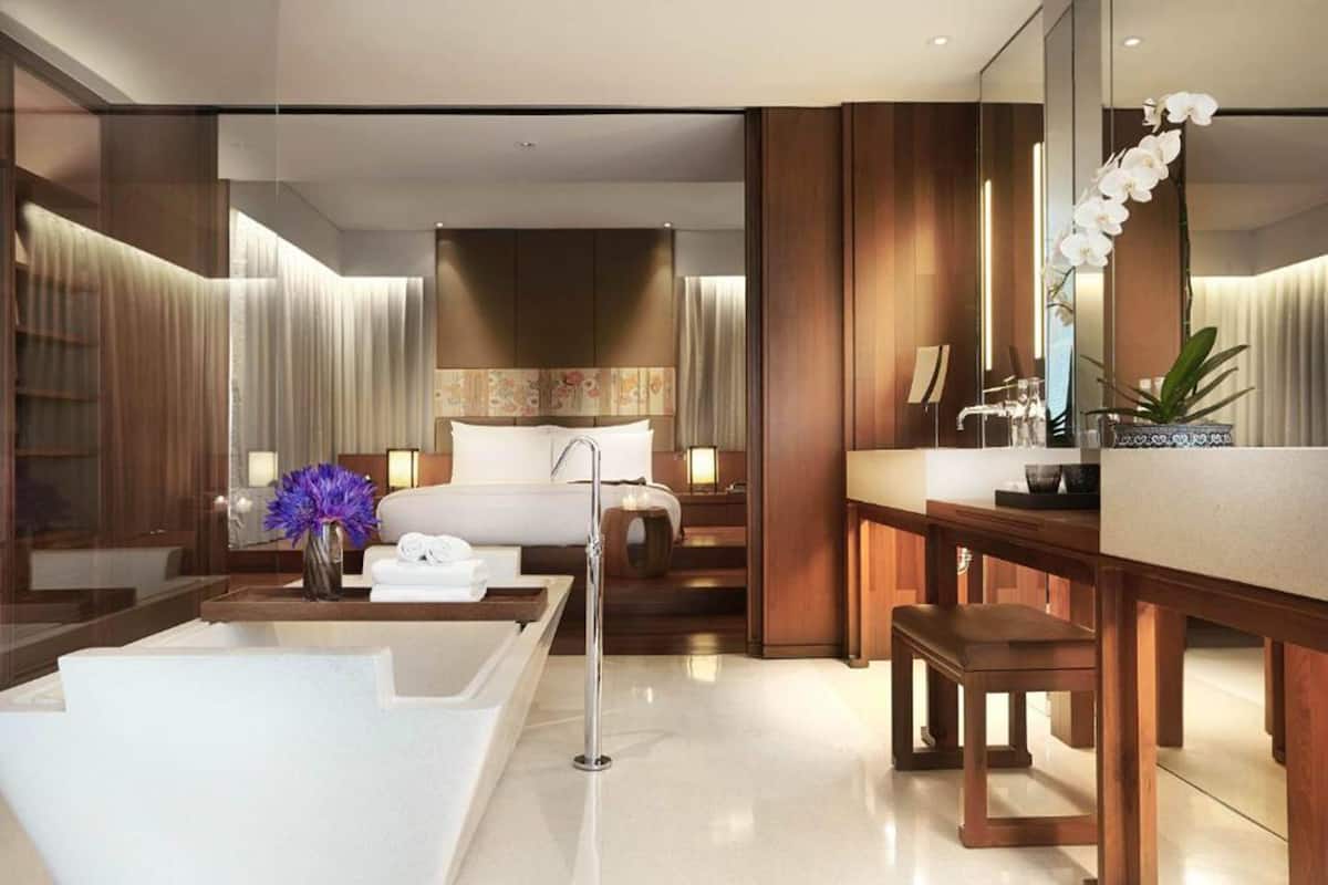 A modern hotel room with a large bed, a separate sitting area with a bench, a bathtub, and a large mirror.