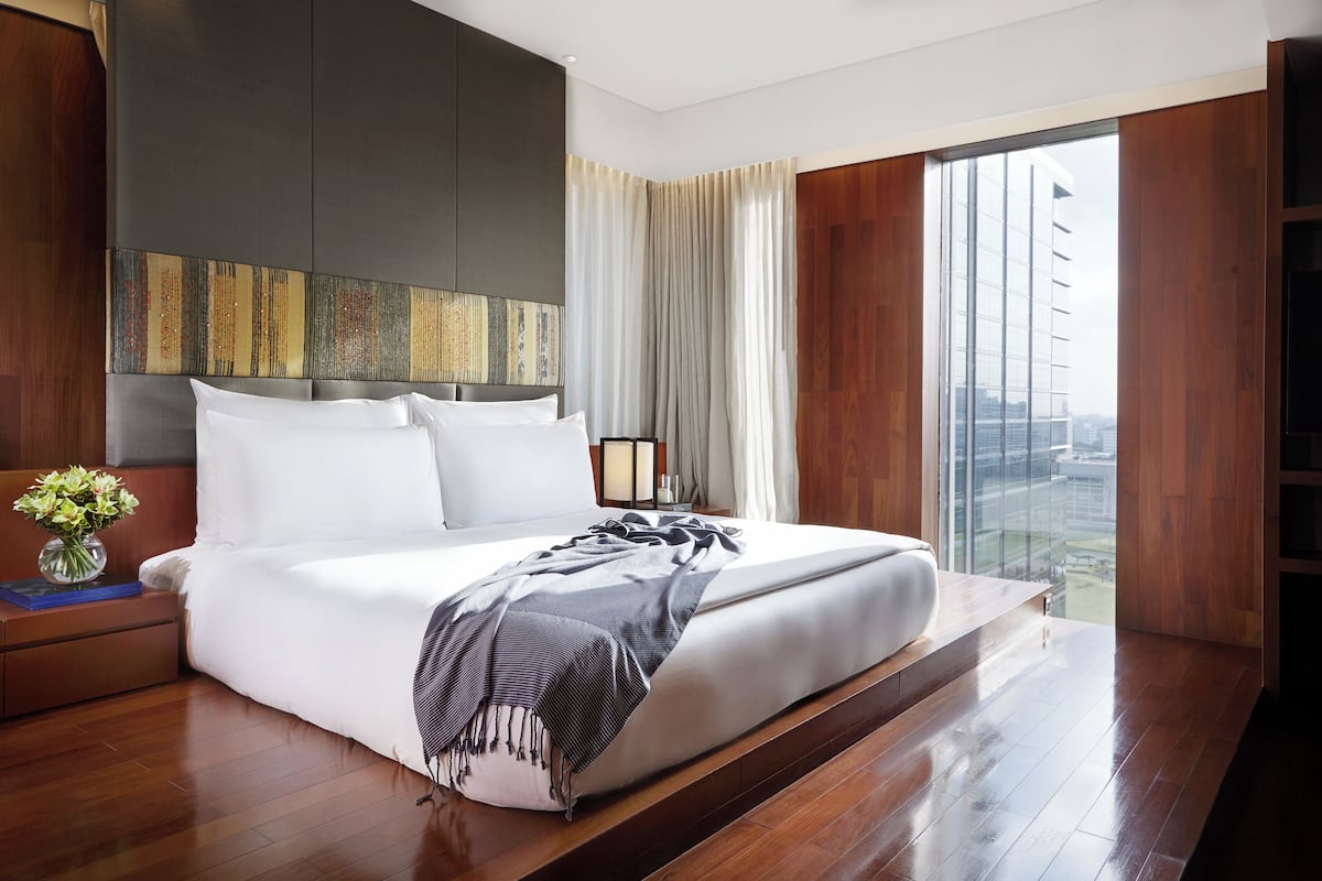 A modern hotel room with a large bed, a bedside table with a vase of flowers, and a view of the city through large windows.