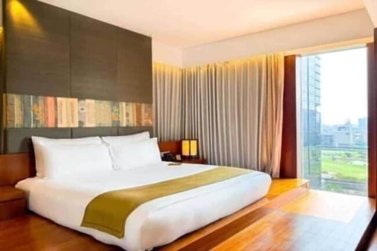 A modern hotel room with a large bed, a nightstand, a lamp, and a view of the city through large windows.