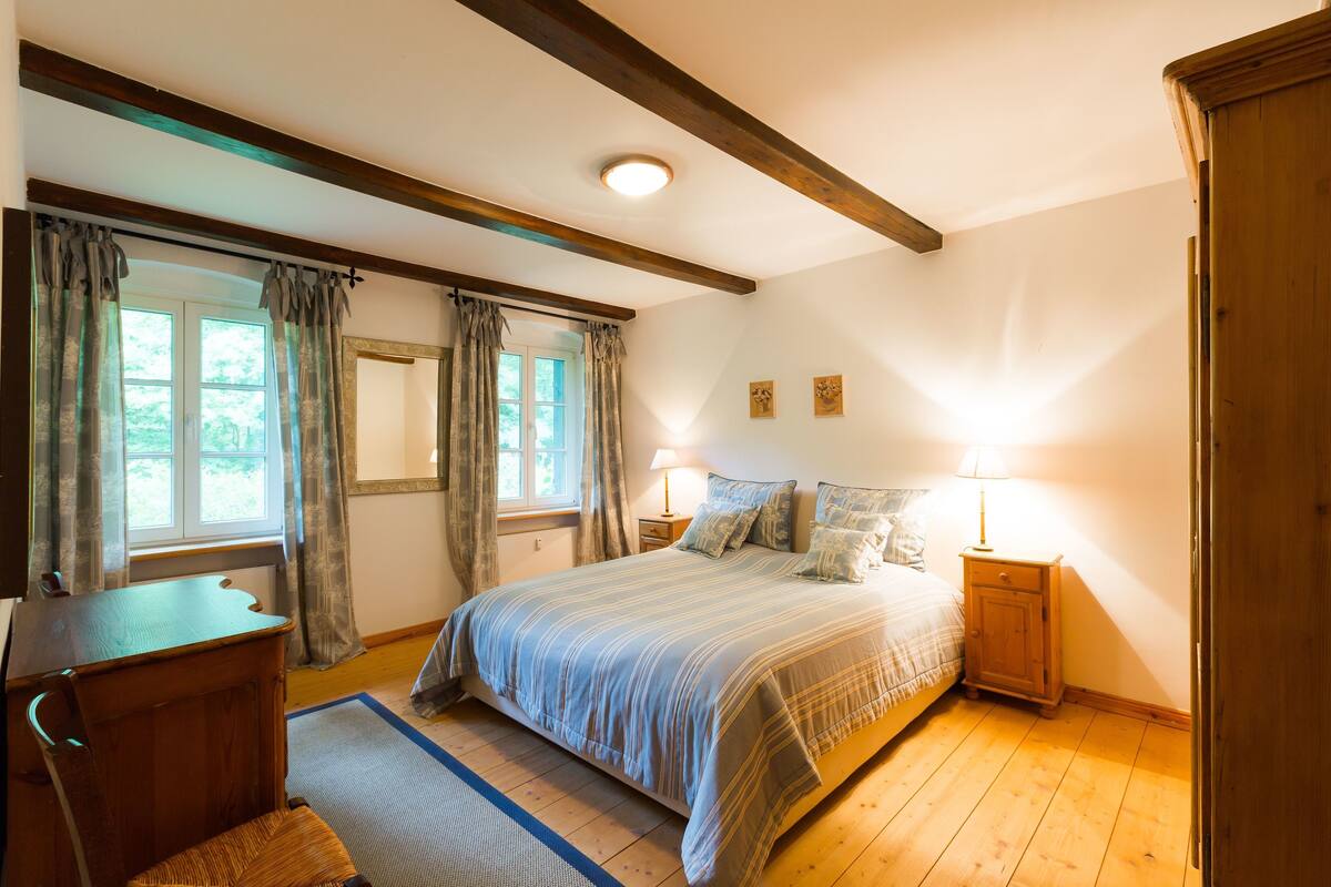 Une chambre avec un lit en bois, un bureau et une chaise. Il y a deux fenêtres avec des rideaux, une table de chevet avec une lampe et une porte.