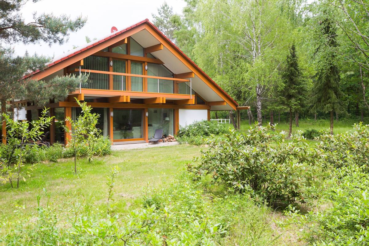 Une maison moderne à deux étages, dotée de grandes fenêtres en verre, d’une terrasse en bois et d’un jardin qui l’entoure.