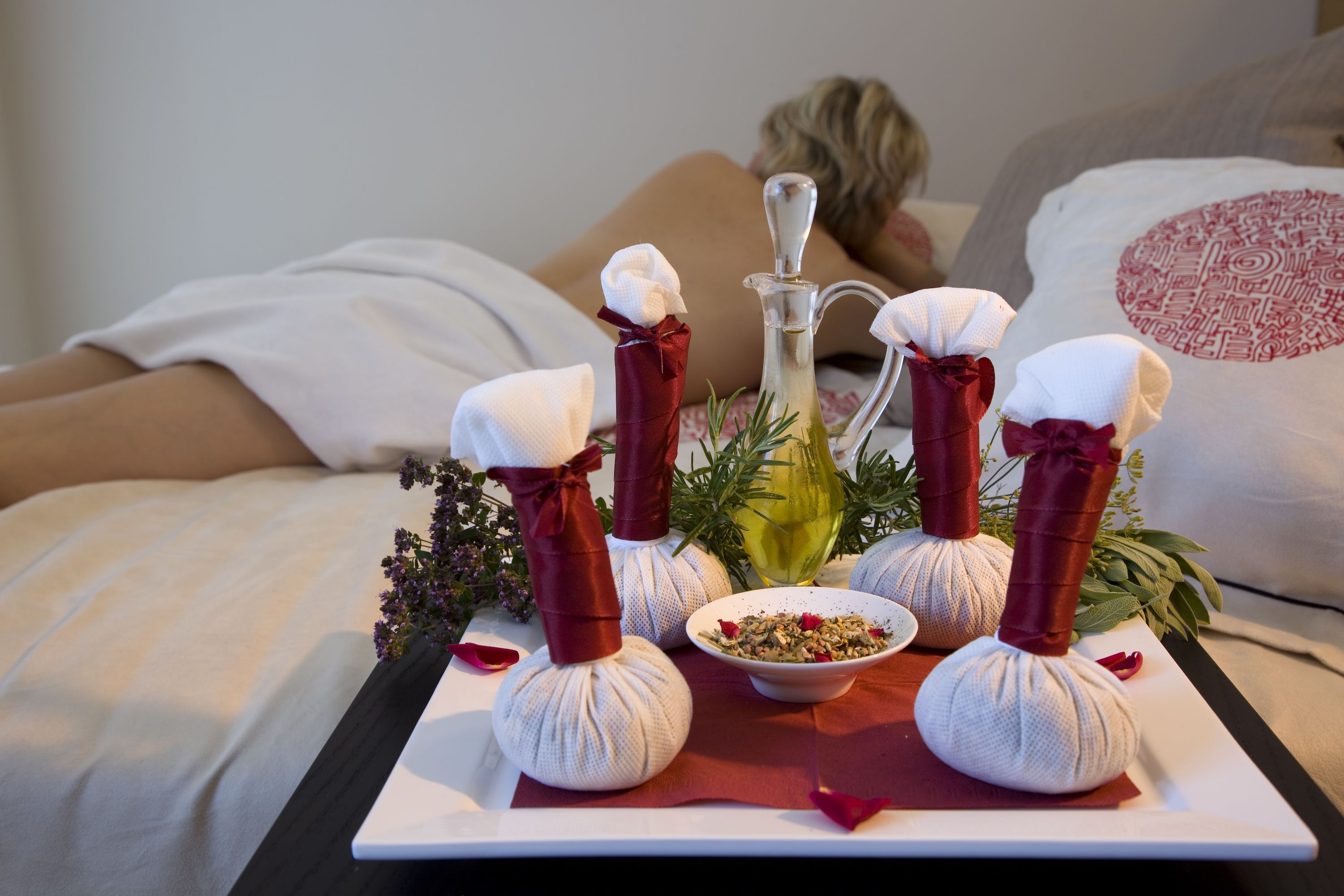 En massageopstilling med håndklæder, en skål med urter og en flaske olie på en bakke.
