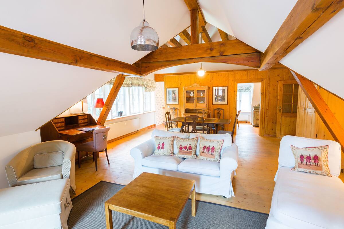 Une pièce mansardée chaleureuse avec un plafond en bois, un ensemble de canapés blancs, une table basse en bois et un coin repas avec une table et des chaises.