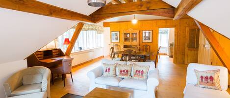 A cozy attic room with a wooden ceiling, a white sofa set, a wooden coffee table, and a dining area with a table and chairs.