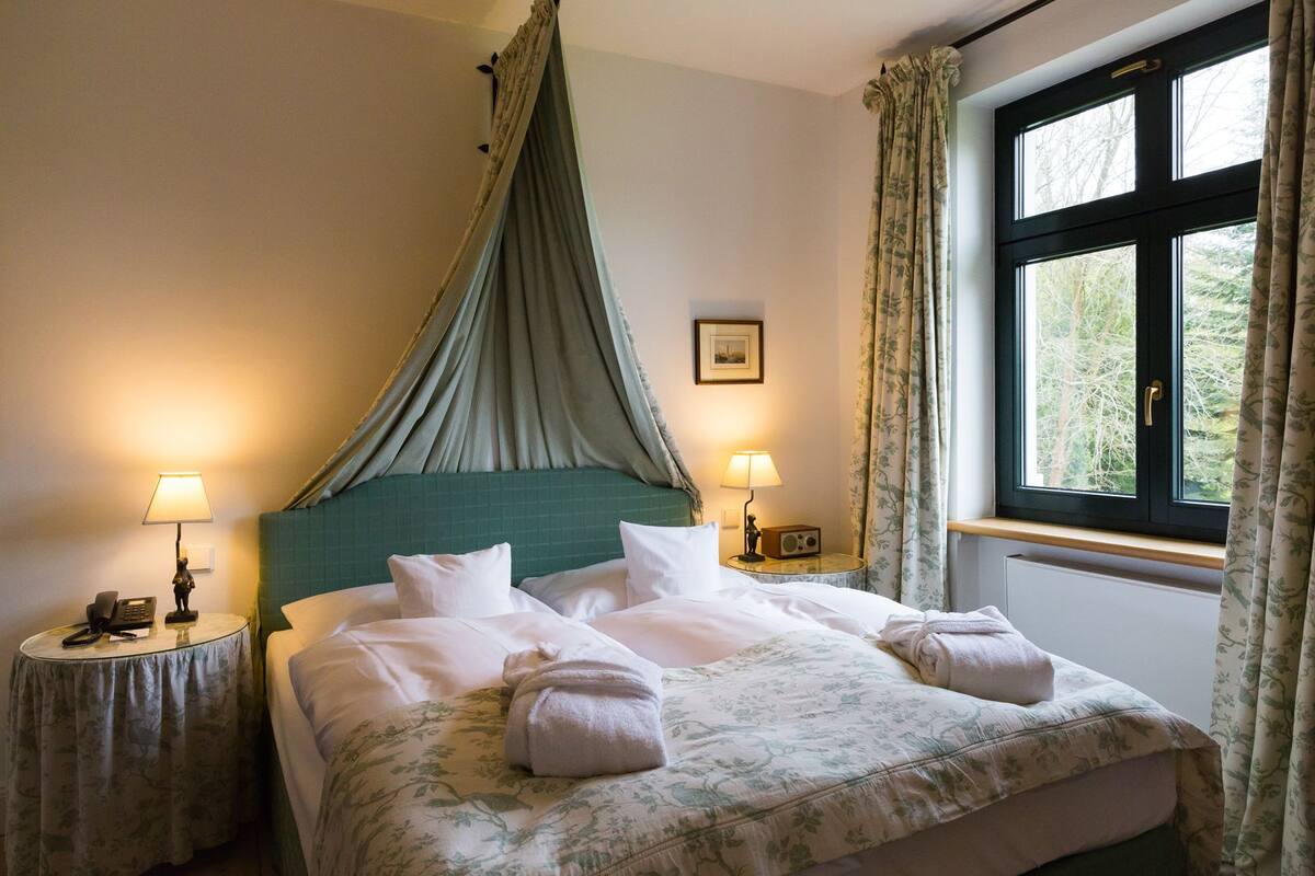 Une chambre d’hôtel avec un lit, deux tables de chevet, une fenêtre avec des rideaux et une vue sur les arbres.