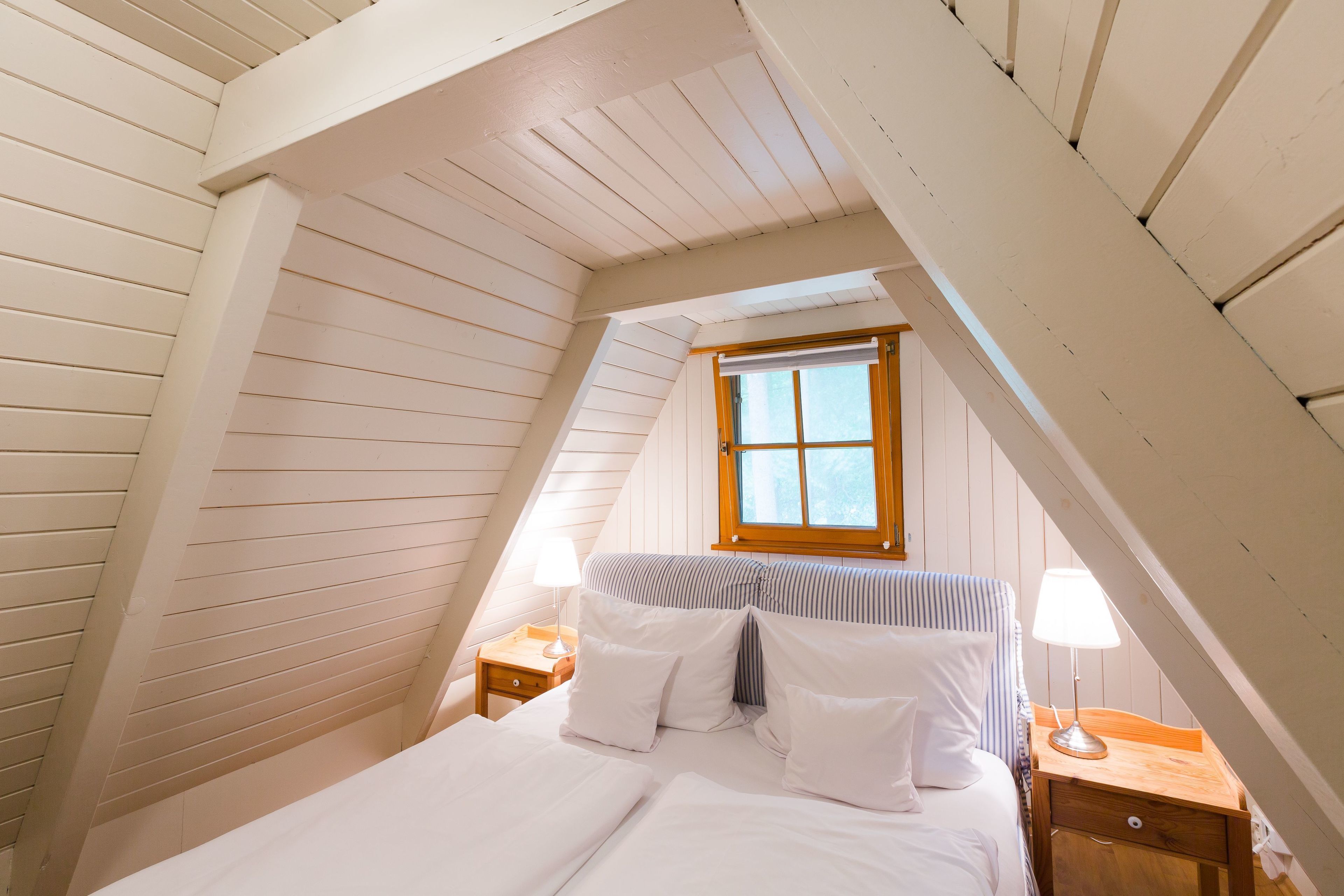 A bedroom with a sloped ceiling, a window, a bed with white bedding, two bedside tables with lamps, and a striped headboard.