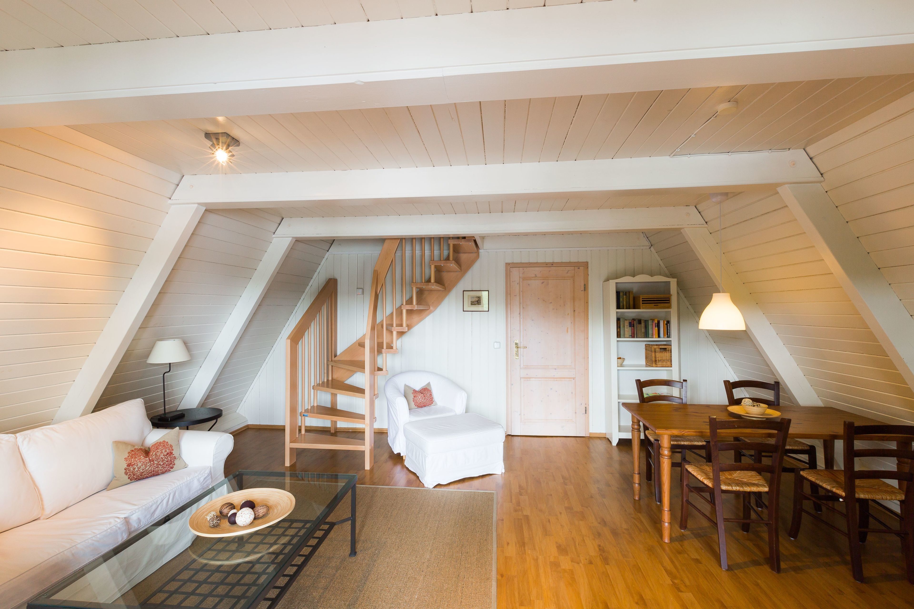 Une chambre confortable située dans les combles, avec un escalier en bois, un lit blanc, une table à manger avec des chaises et une bibliothèque.