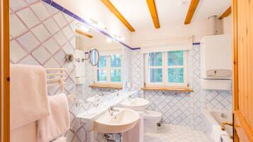 Un bagno con tre lavandini, un bidet e una vasca da bagno. Asciugamani appesi a un porta asciugamani, e una finestra lascia entrare luce naturale.
