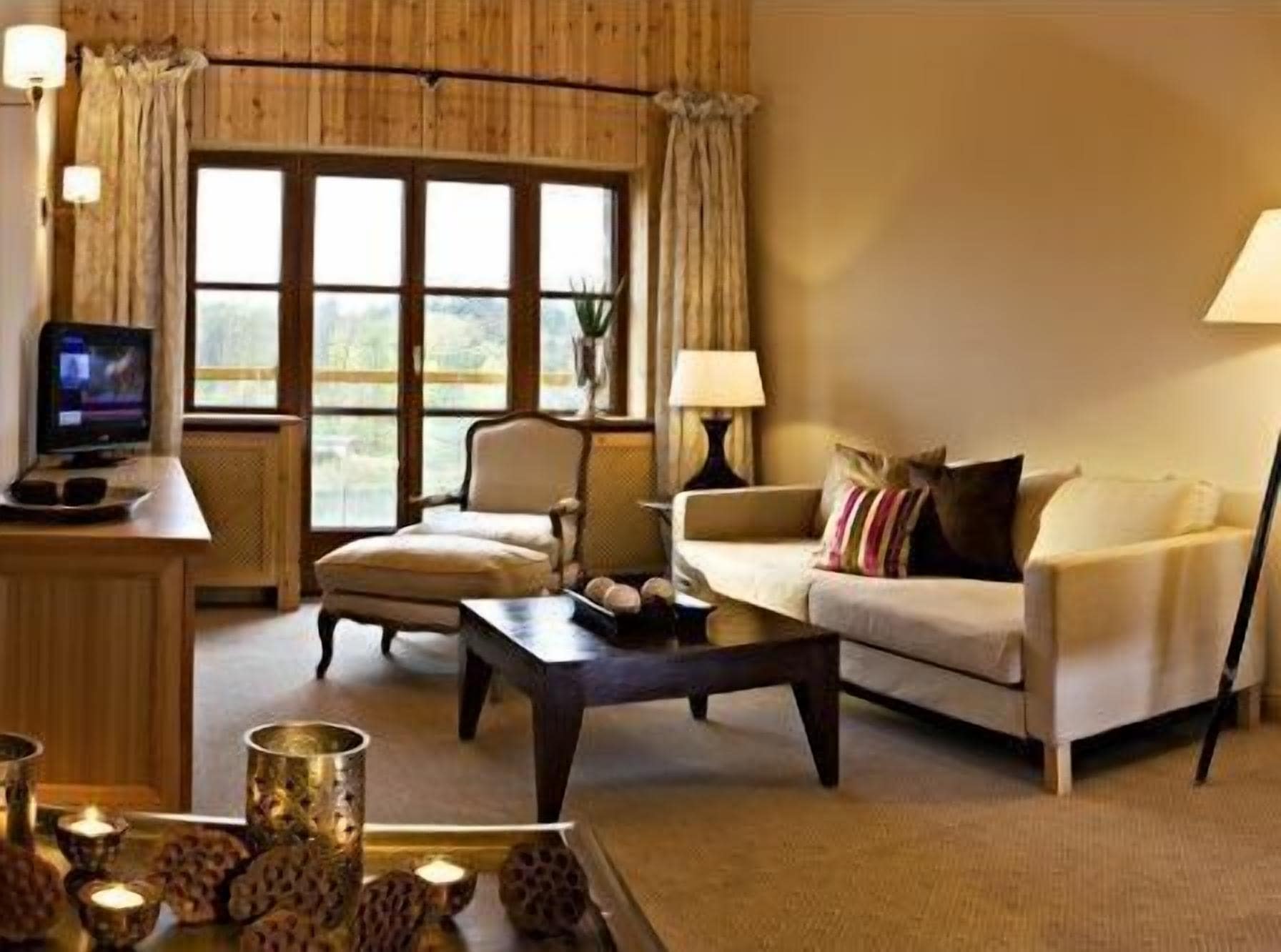Una sala de estar acogedora con un sofá, un sillón y una mesa de centro. Hay un televisor sobre una mesita de noche de madera, y una lámpara sobre otra mesita. La habitación tiene ventanas grandes con cortinas.