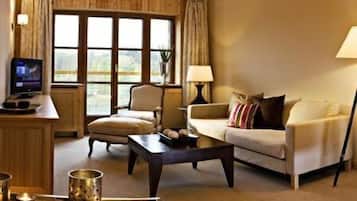 Una sala de estar acogedora con un sofá, un sillón y una mesa de centro. Hay un televisor sobre una mesita de noche de madera, y una lámpara sobre otra mesita. La habitación tiene ventanas grandes con cortinas.