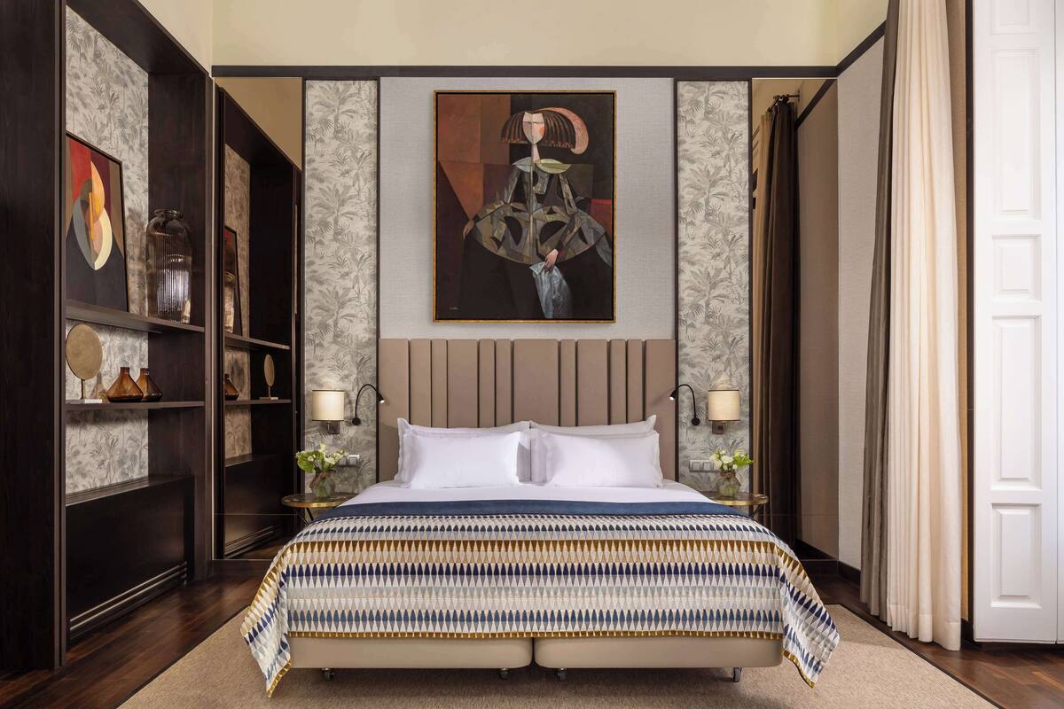 A bedroom with a large bed, a headboard with vertical slats, a wall-mounted painting, and a nightstand with a lamp.