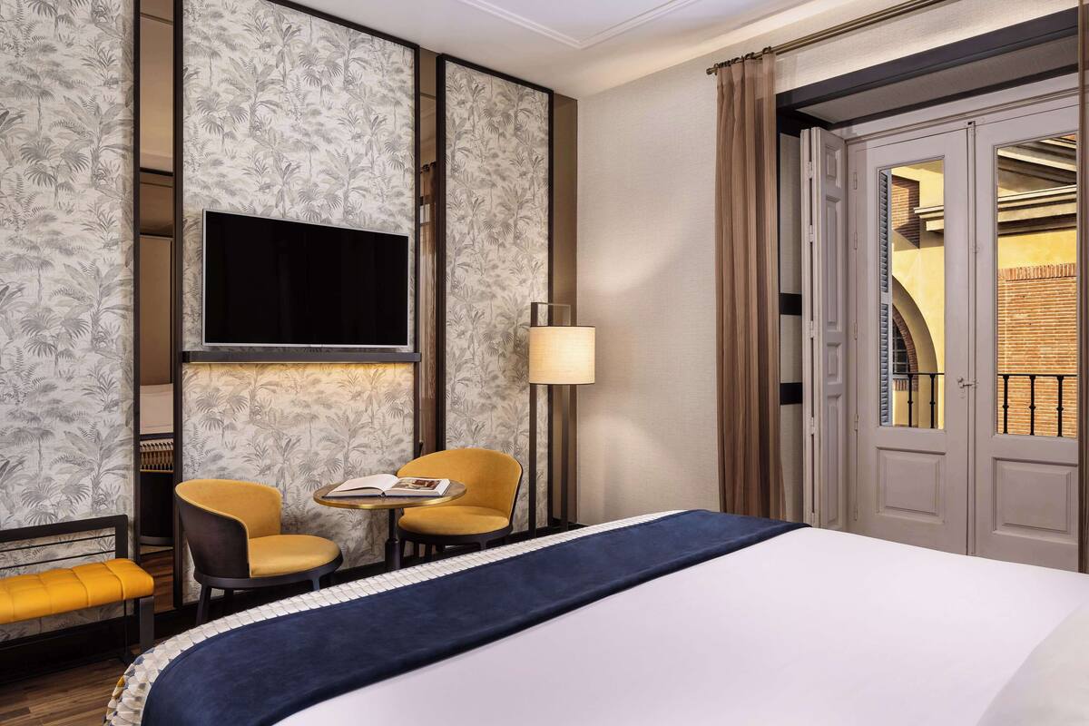 A hotel room with a bed, two chairs, a small table, a TV, and a door leading to a balcony.
