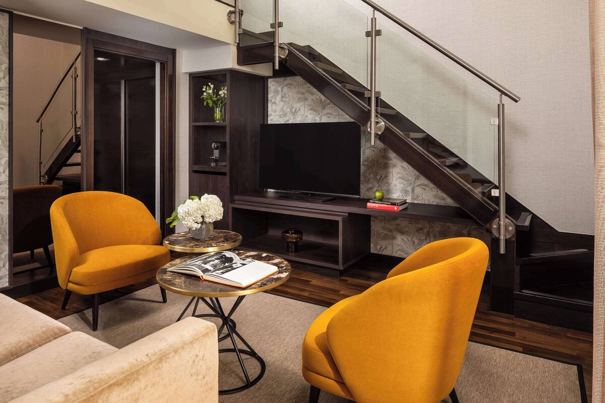 A modern living room with two yellow armchairs, a flat-screen TV, a bookshelf, and a staircase.