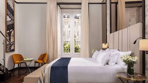 A hotel room with a bed, two chairs, a small table, a vase of flowers, and a view of the city through a door. - NH Collection Palacio de Tepa (Madrid)