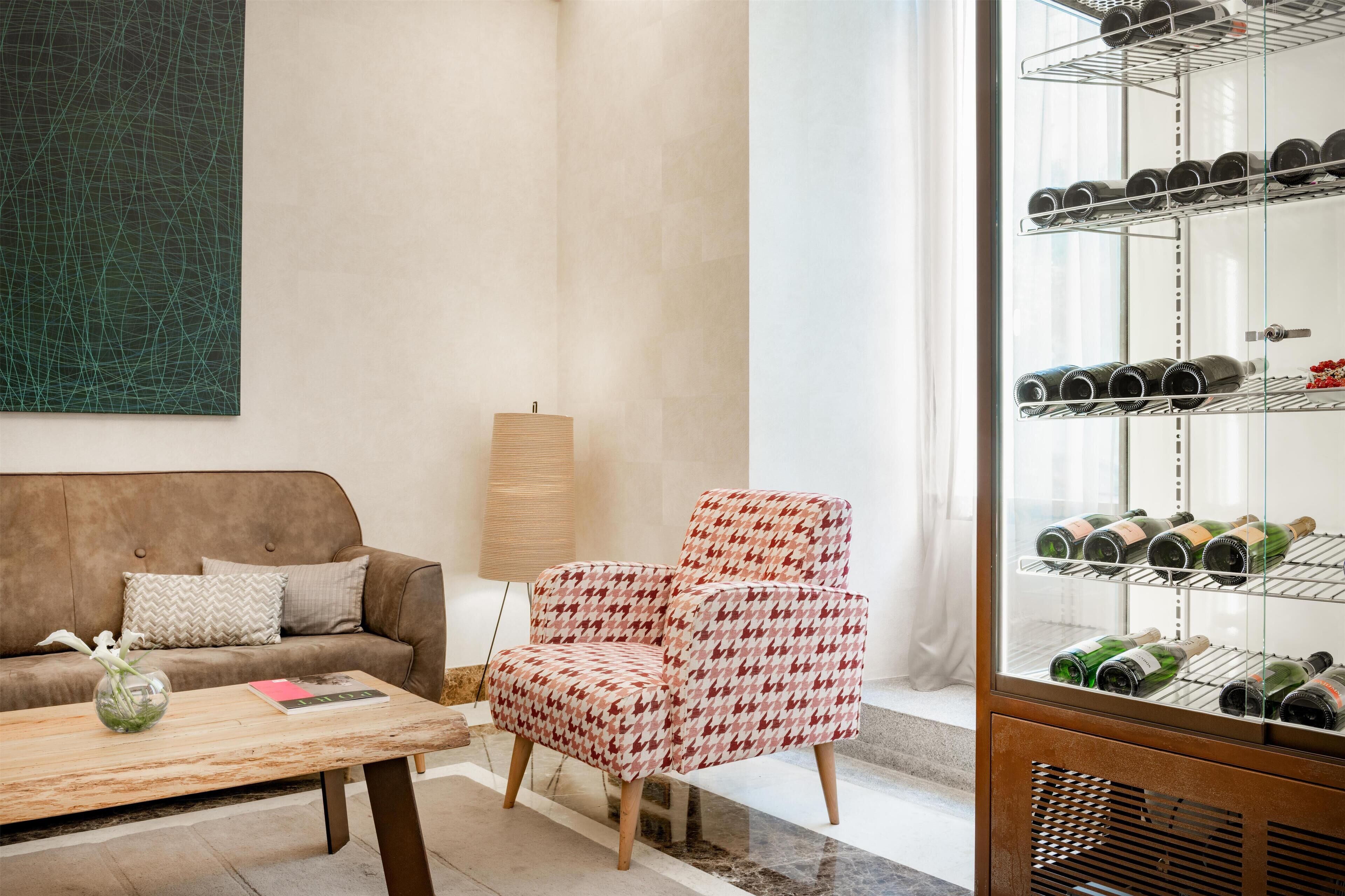Ein Wohnzimmer mit einem braunen Sofa, einem gemusterten Sessel, einem Weinkühlschrank aus Glas und einem Couchtisch mit Vase und Buch.