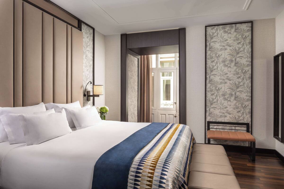 A hotel room with a large bed, a bench, a desk, and a view of the outside.
