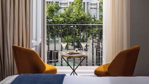 A hotel room with a bed, two yellow chairs, a small round table with a plate of food and two glasses, and a balcony with a view of buildings and trees. - NH Collection Palacio de Tepa (Madrid)