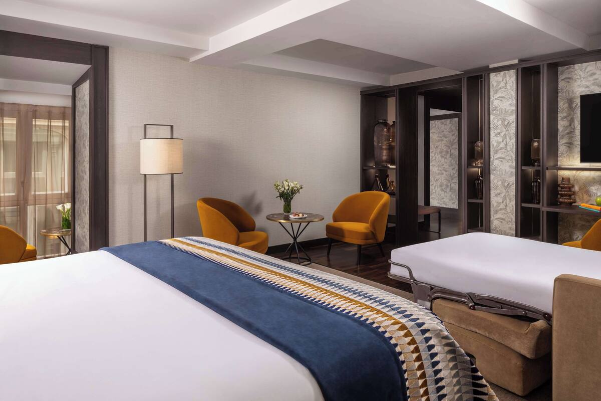 A hotel room with a large bed, two yellow armchairs, a small round table with a vase, and a television on the wall.