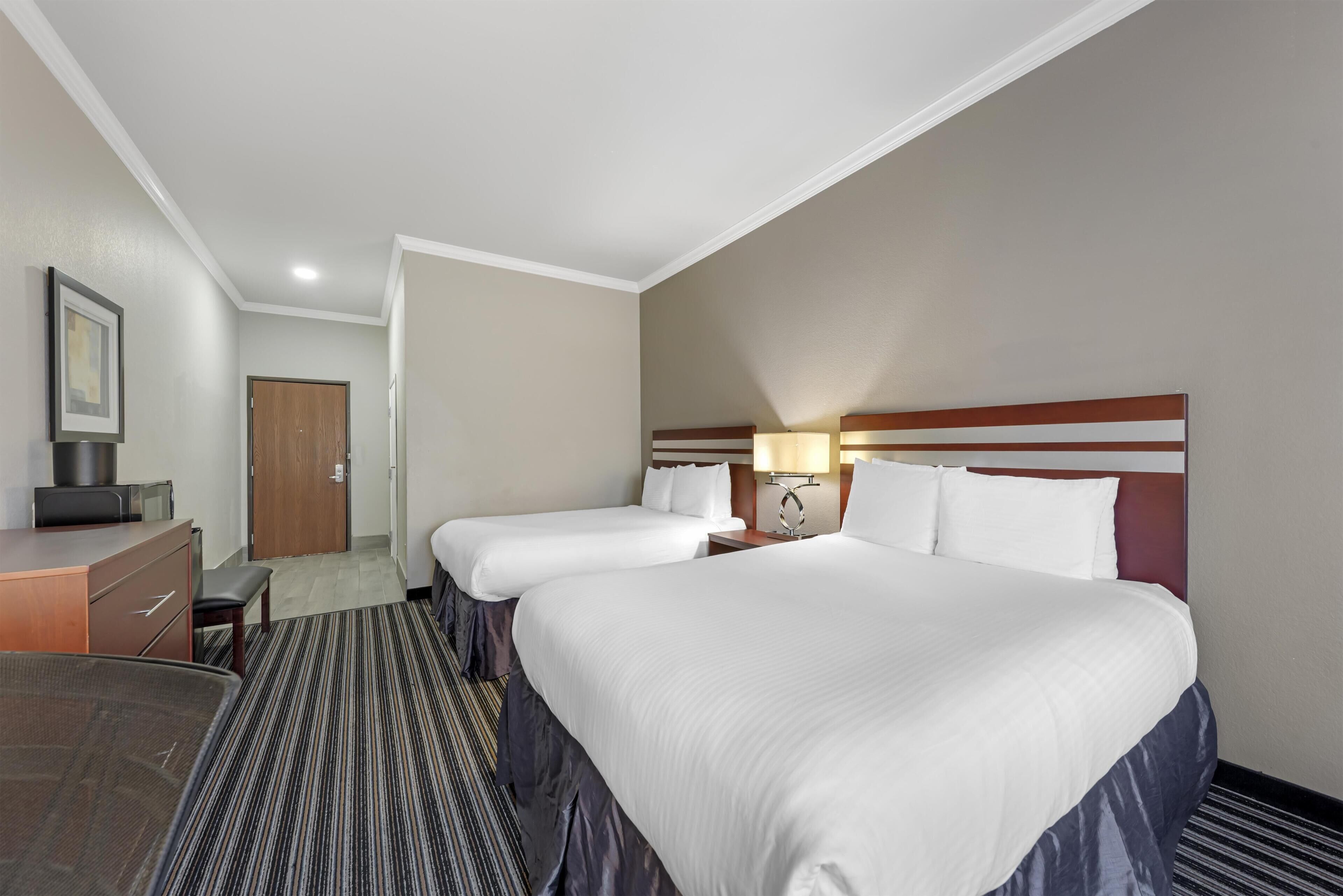 A hotel room with two beds, a desk, a chair, and a door.