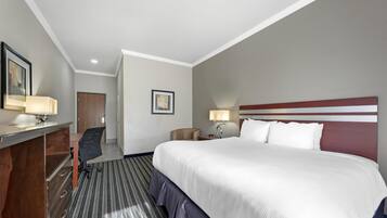 A hotel room with a large bed, a desk with a chair, a desk lamp, a bedside table with a lamp, and a door.
