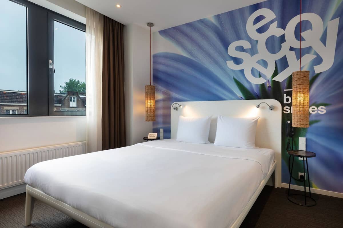 A hotel room with a large bed, a view of the city through a window, a wall with a large floral design, and a wicker lamp.