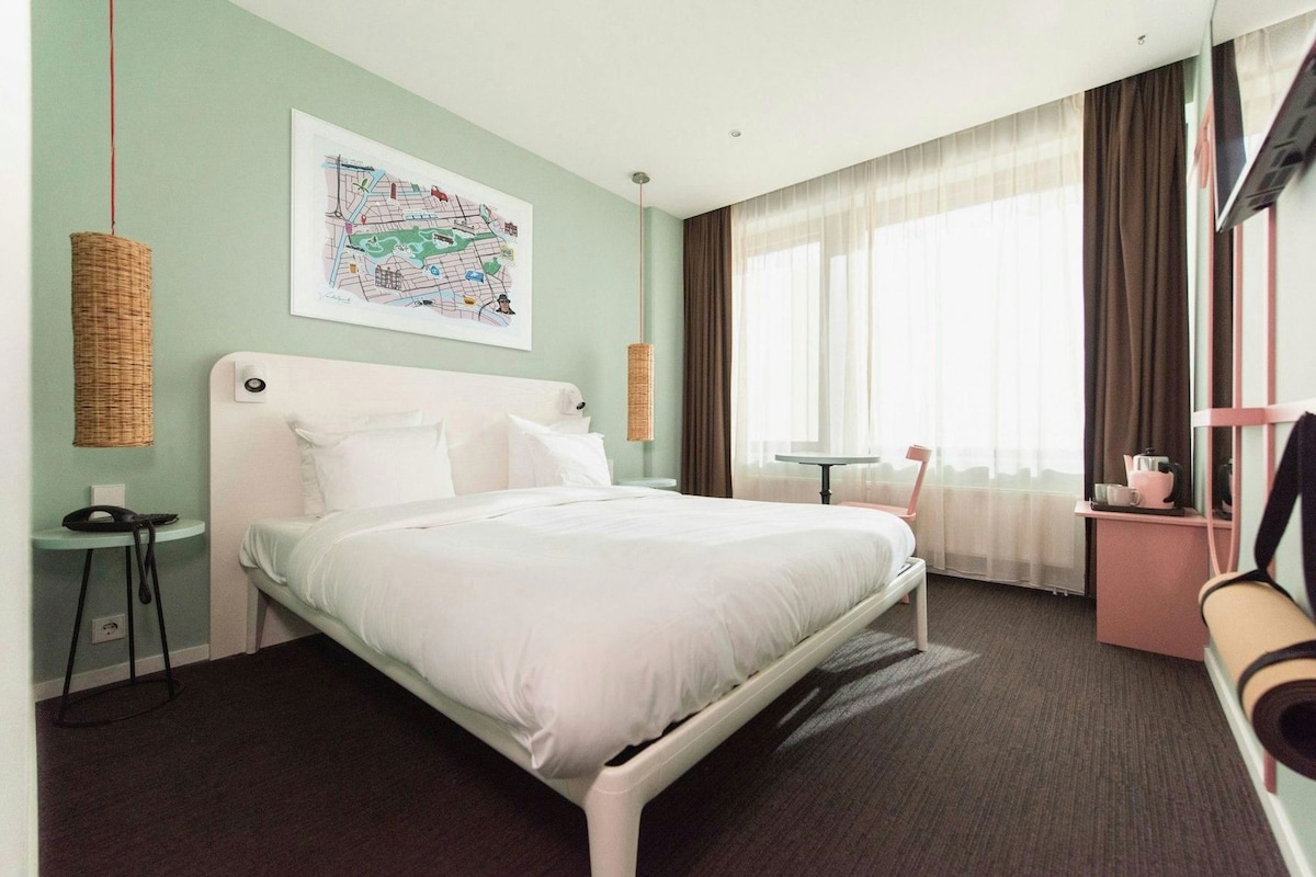 A hotel room with a bed, a desk, a chair, a map on the wall, and a window with curtains.