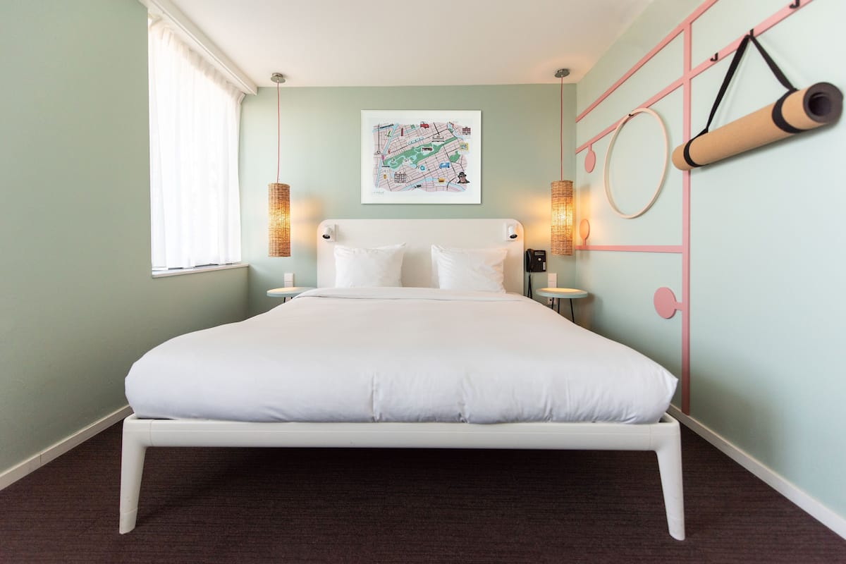 A neatly made bed in a room with a light green wall, a map artwork, and a yoga mat hanging on the wall.