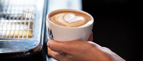 A person holding a cup of coffee with a heart-shaped latte art, with a coffee machine in the background.