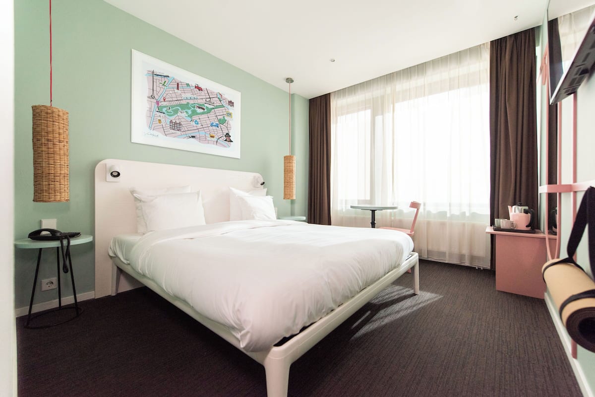 A hotel room with a bed, a small table, a chair, a map on the wall, and a window with curtains.