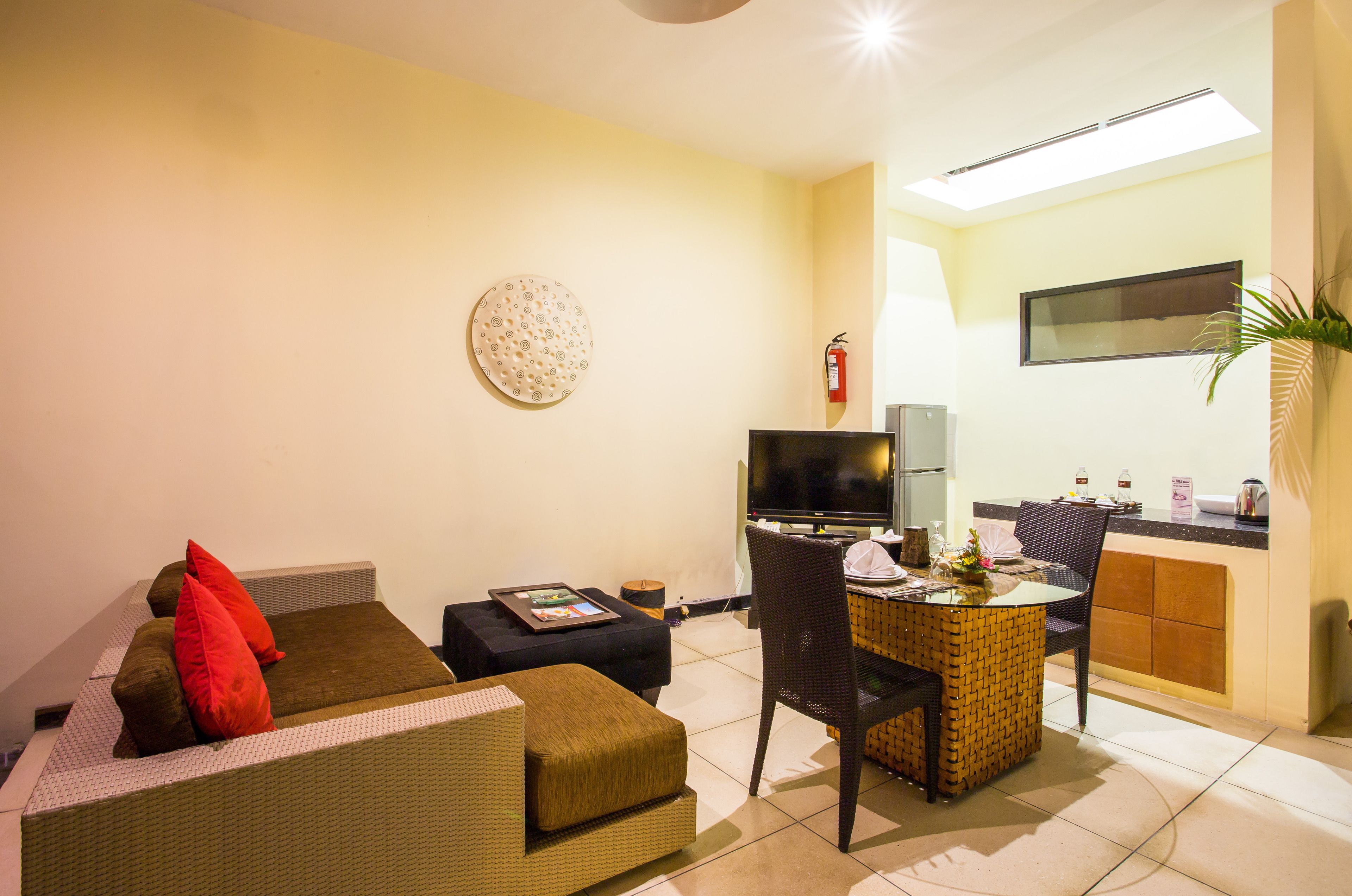 A compact living room with a sofa, a small table, and a TV. A kitchen area is visible in the background with a flat-screen TV and a fire extinguisher.