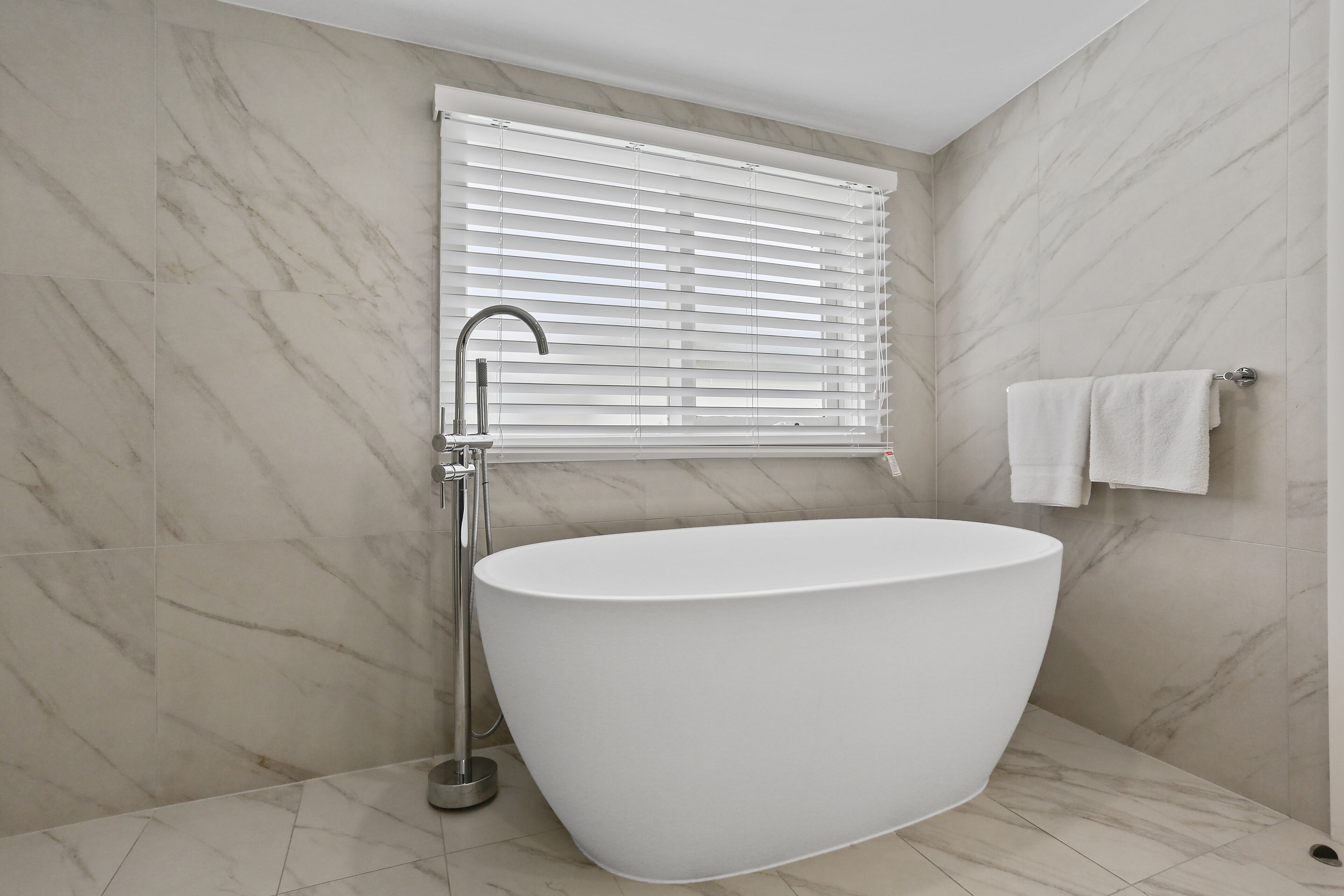 A modern bathroom with a freestanding bathtub, a showerhead, and a window with blinds.