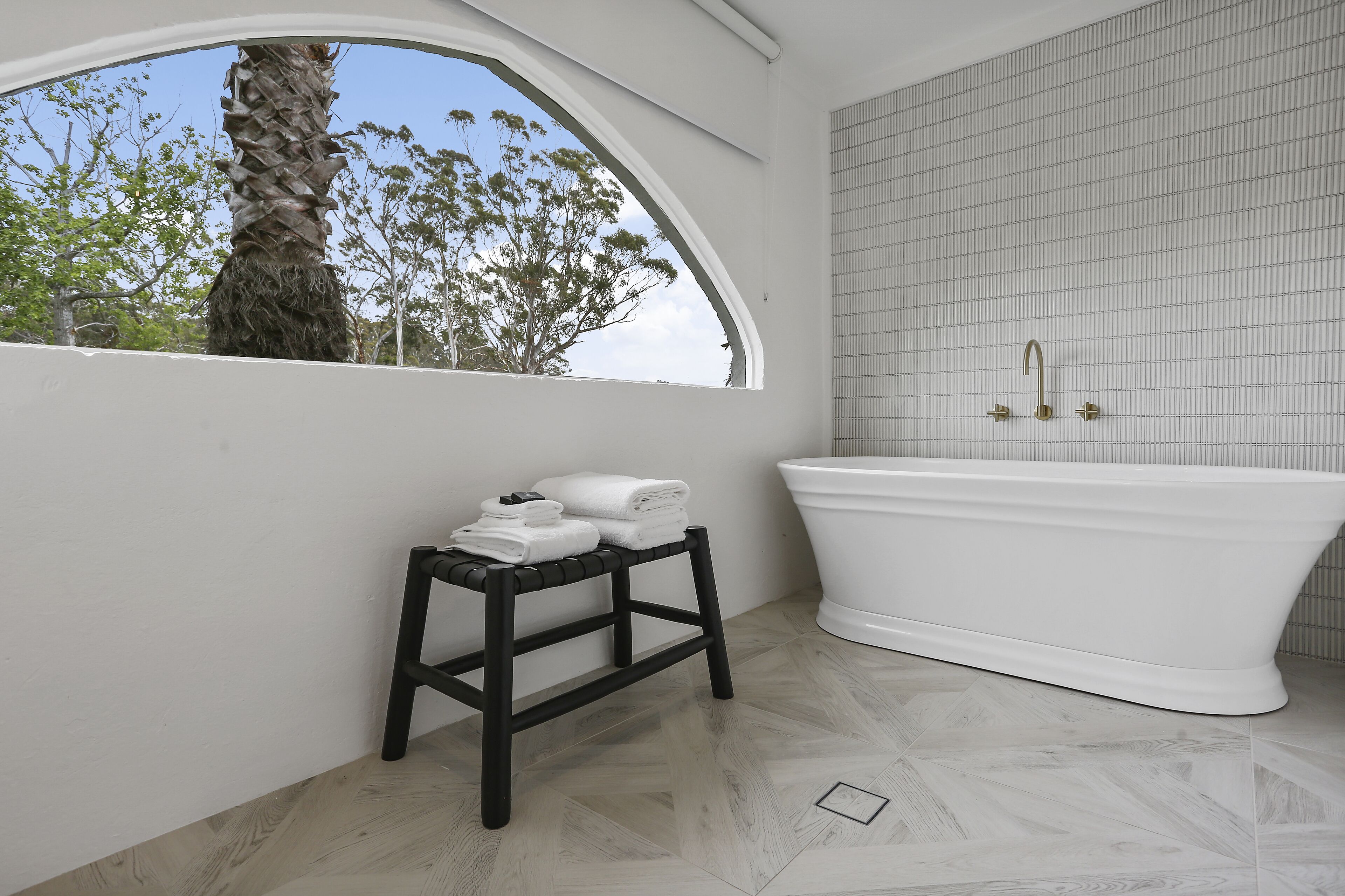 Un baño con una bañera independiente, un taburete pequeño con toallas y una ventana con vistas a árboles.