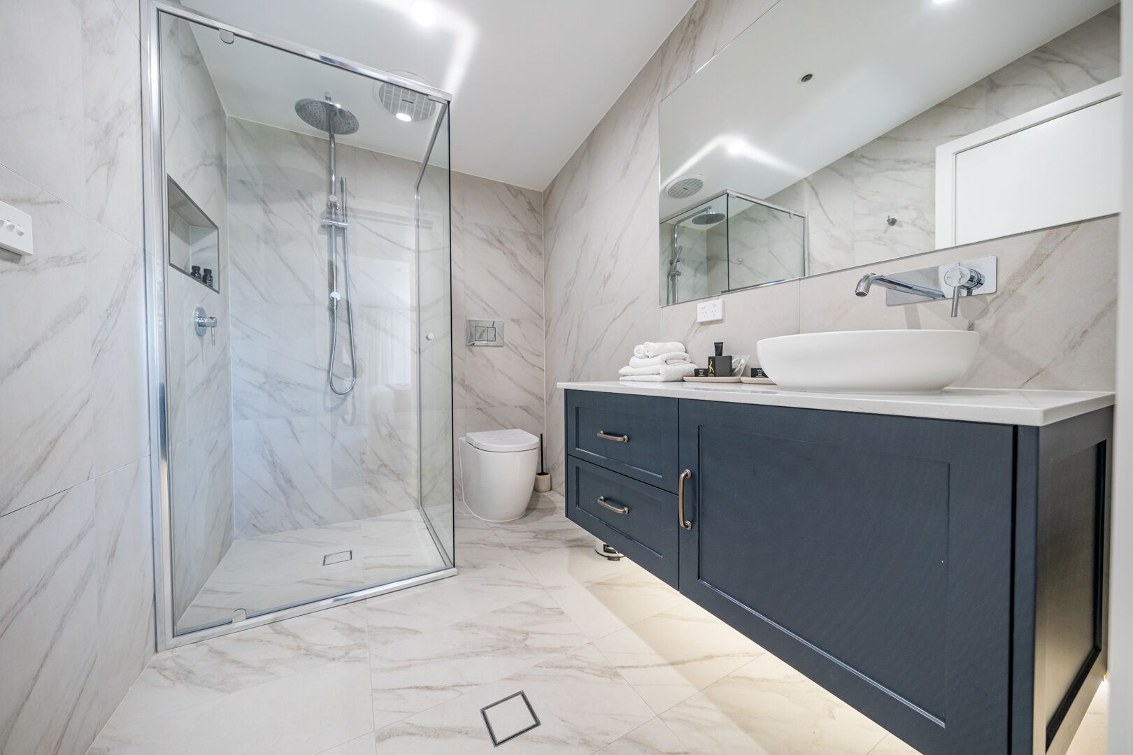 A modern bathroom with a glass-enclosed shower, a white sink, dark cabinets, and marble flooring.