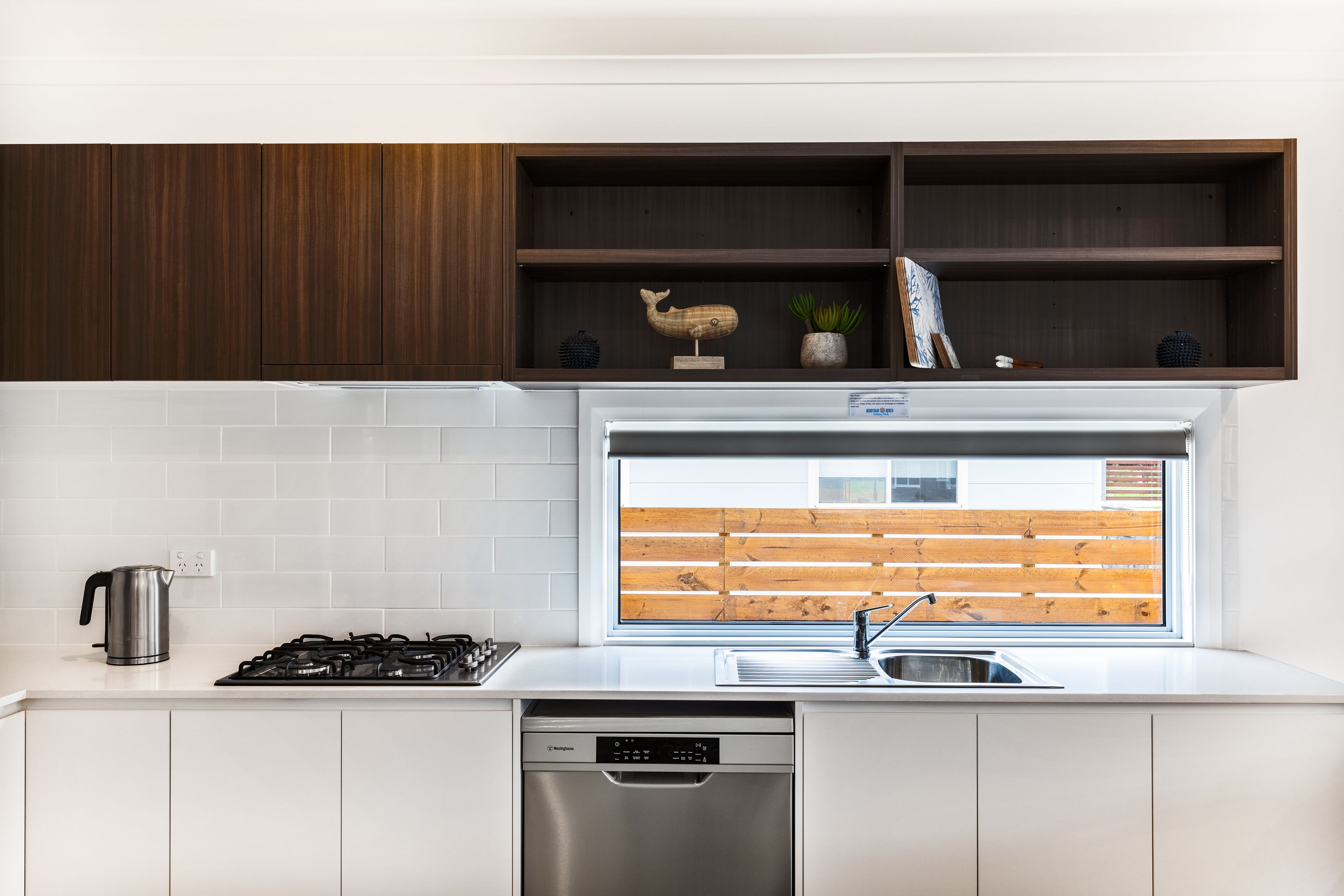 A modern kitchen with white cabinets, a stainless steel stove, a dishwasher, and a window with a view of a wooden deck.